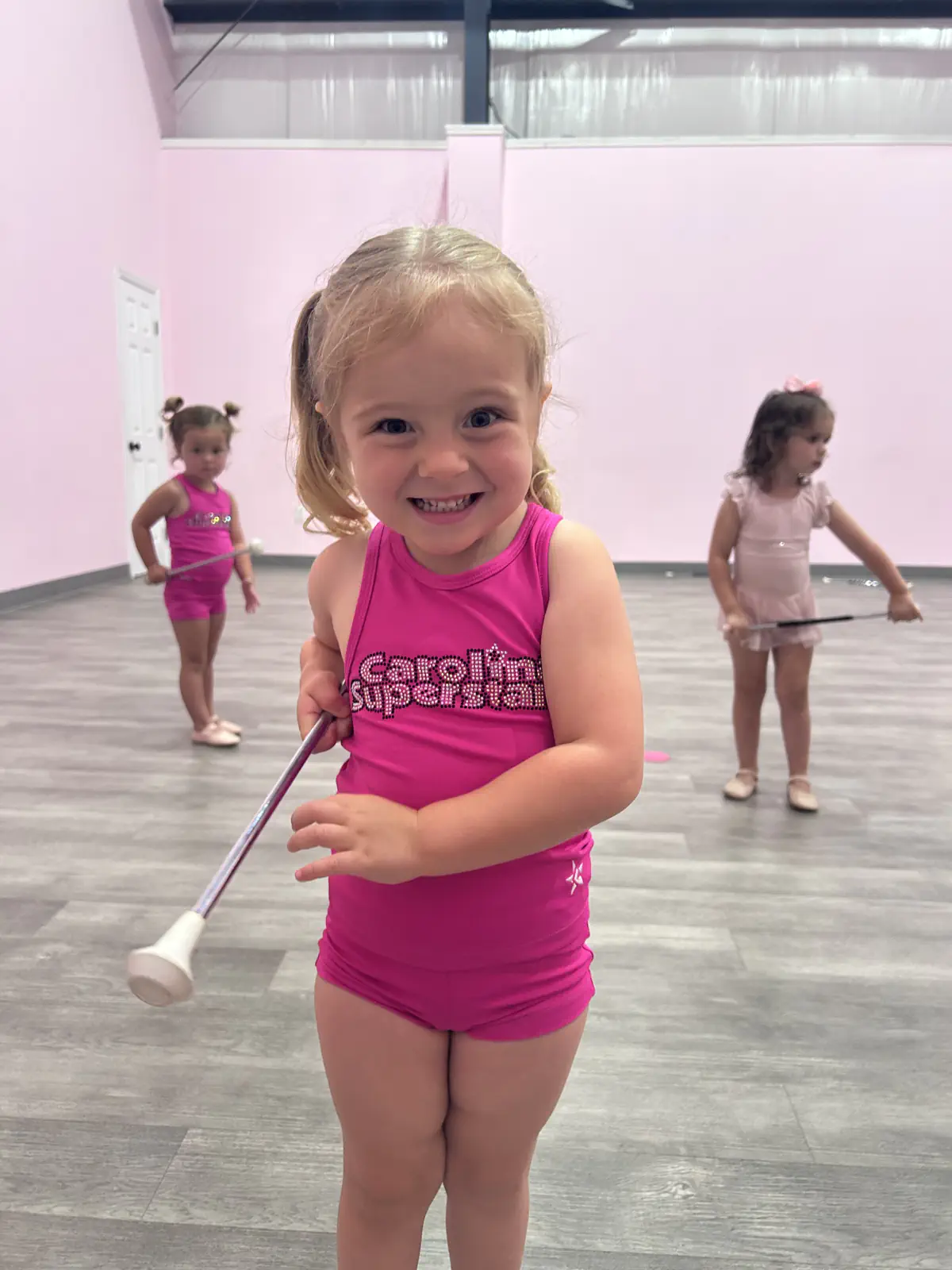 Smiling toddler girl in pink leotard holding baton at dance class