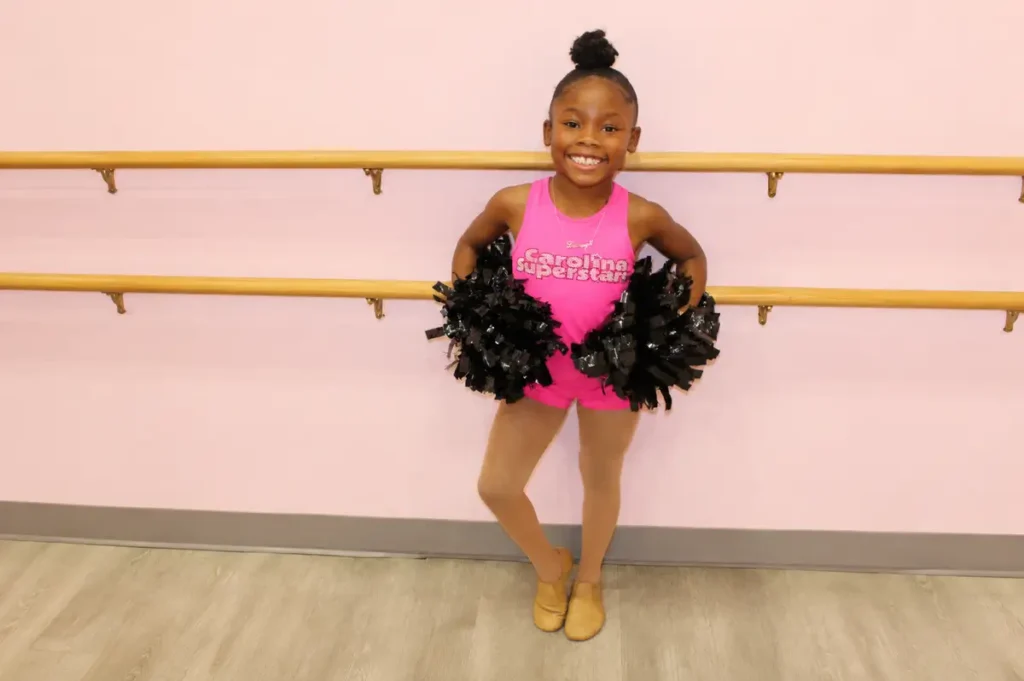 Young girl in pink Carolina Superstars outfit holding black pom-poms
